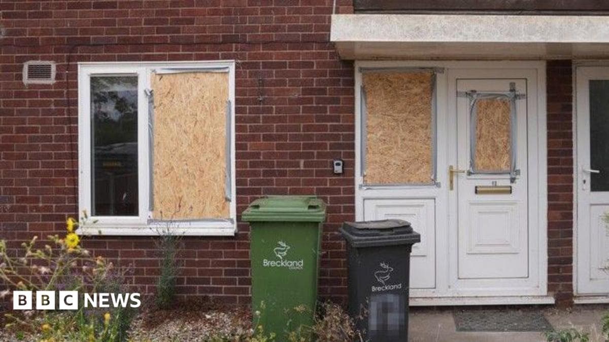 The front of house where several windows have been boarded up. The garden is untidy. In front of the property are two bins, one green and the other black.
