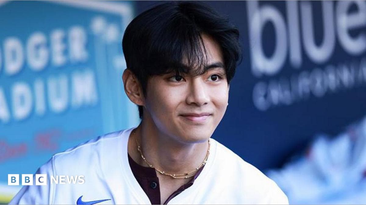 V smiles as he looks at a distance, wearing a baseball shirt