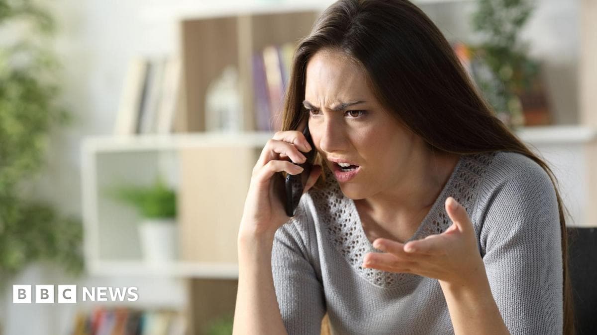 Woman with angry expression holds phone up to her ear looking frustrated. She has brown hair and wears a grey knit top.