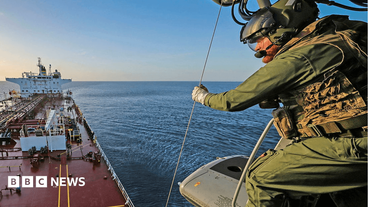 A still showing a military personnel on a helicopter above a large vessel. He is wearing green tactical gear and a helmet and gloves and is looking down at the boat. The water and sky are blue.