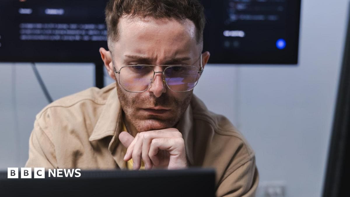 A software engineer, wearing a light brown shirt, looks at a computer monitor in front of him with a concerned expression. A larger screen with computer code on it is on a wall behind him.