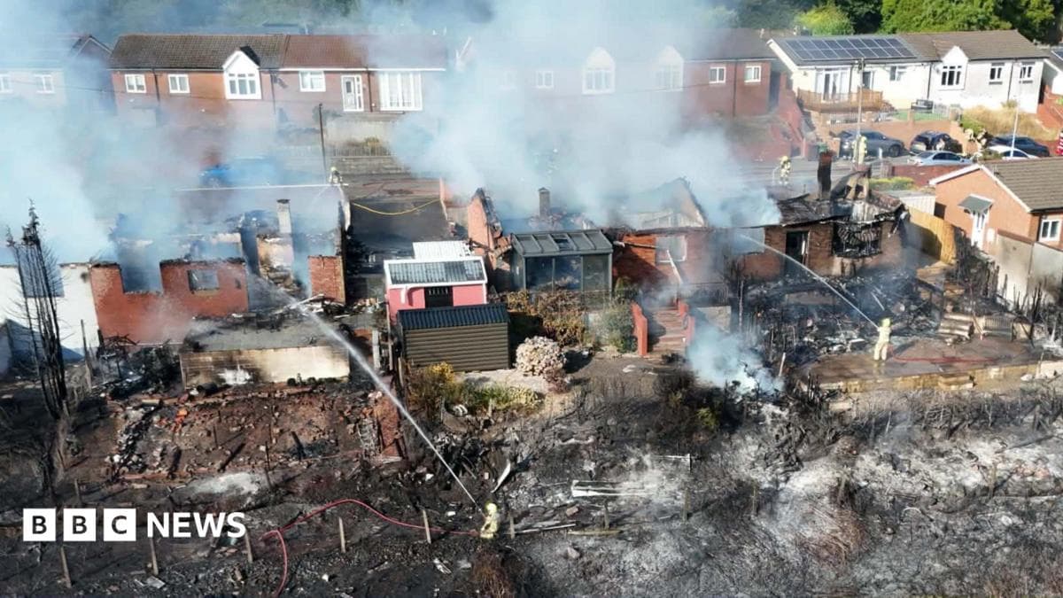 An aerial shot of a row of homes with their roofs destroyed by fire. Smoke streams into the air. Land in front of the houses is scorched, with firefighters holding hoses spraying water towards the scene.