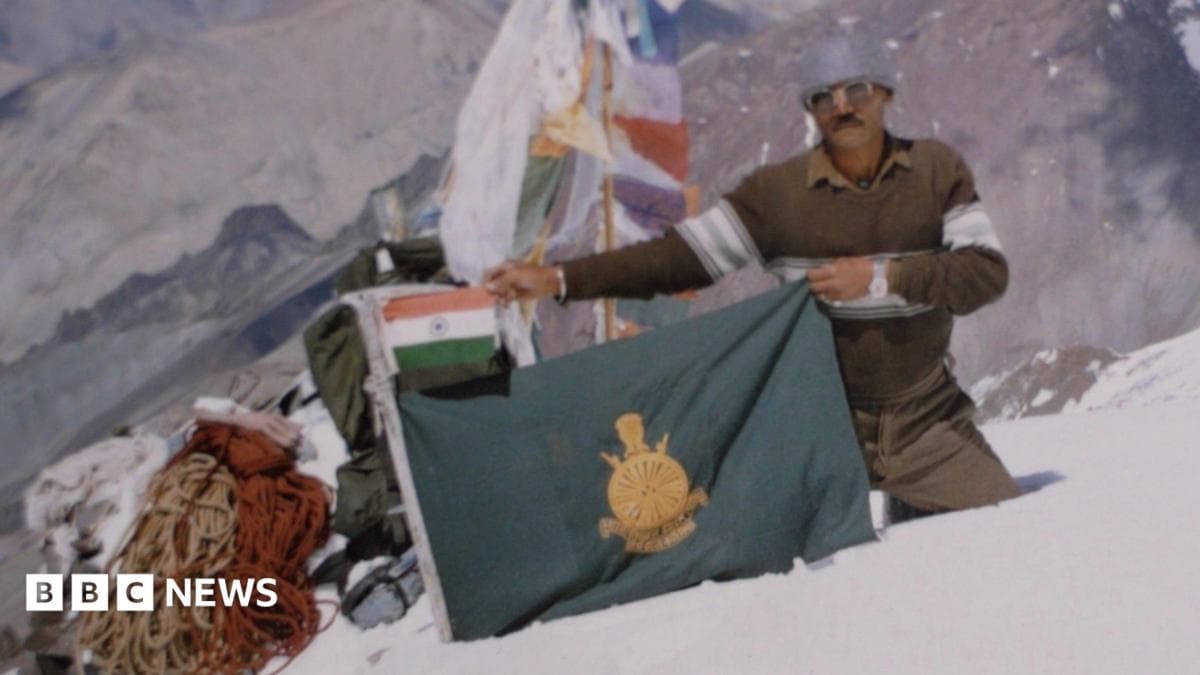 A man wearing protective clothing on a snow-capped mountain peak holds a green flag, with a smaller Indian flag above it.