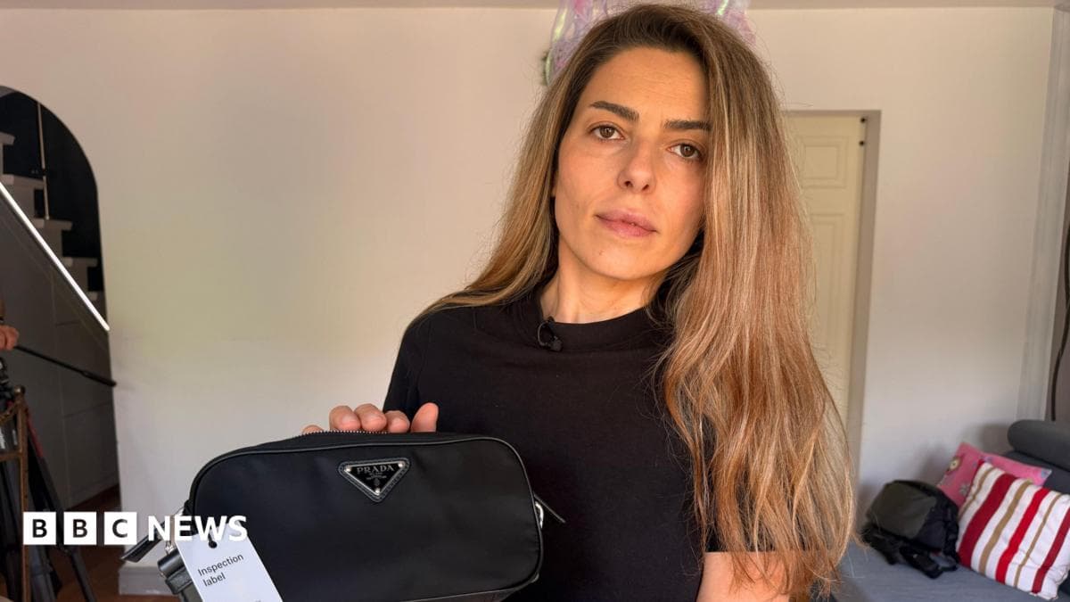 A woman with long dark blond hair holding a black handbag with the Prada logo on it which is fake. She is standing in a living room in a house. It has a grey sofa and multi-coloured cushions. An open staircase can be seen in the background too. She looks serious.