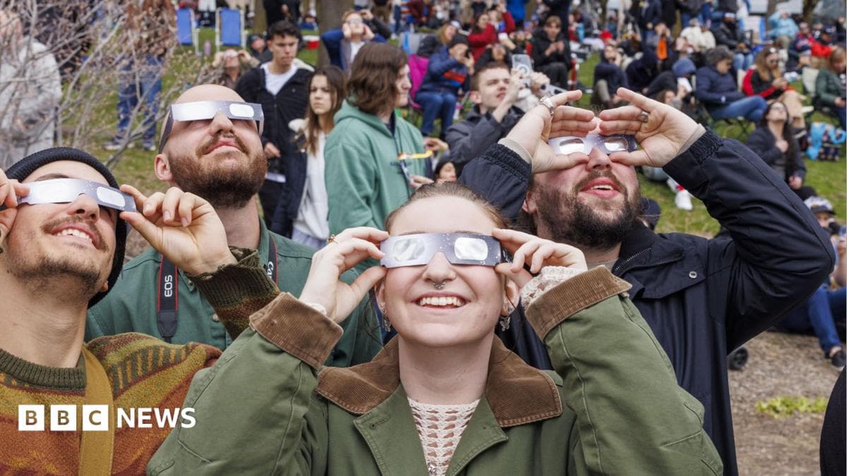 A group of people all holding special glasses to their faces and looking up at the sky to view a solar eclipse. They are wearing coats and in the background are crowds of people standing or sitting on a patch of grass.