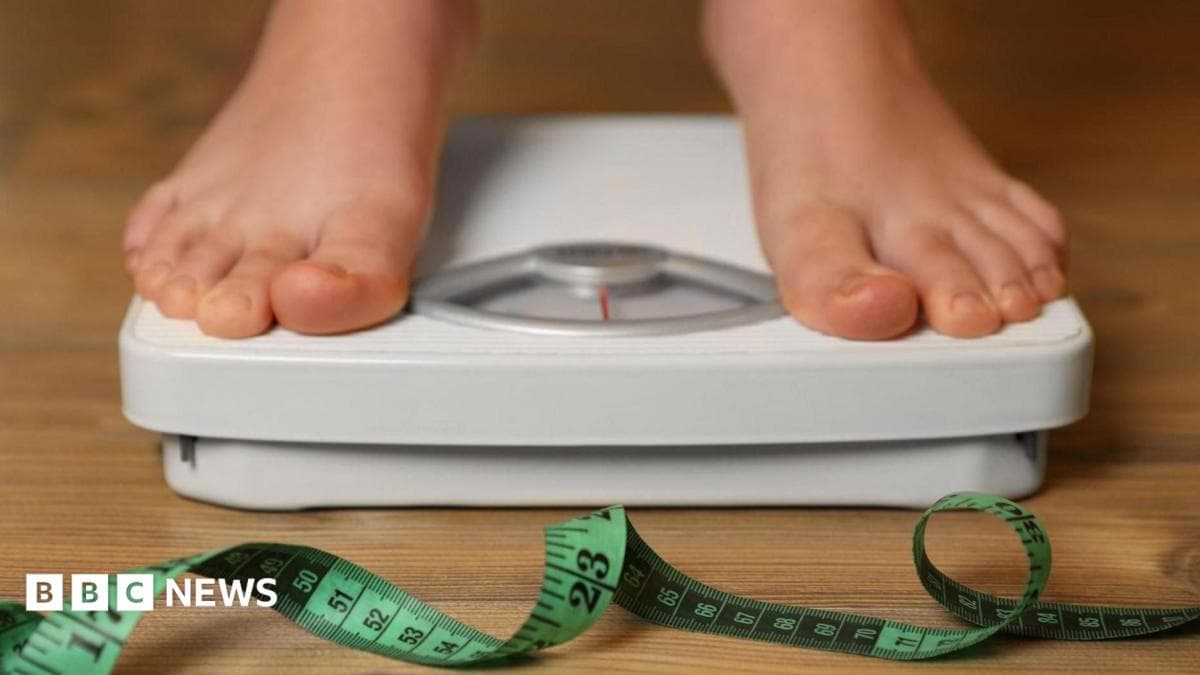 Person on scales in bare feet. The scales are white with a red needle and there is a green fabric tape measure on the wooden floor in front of them.