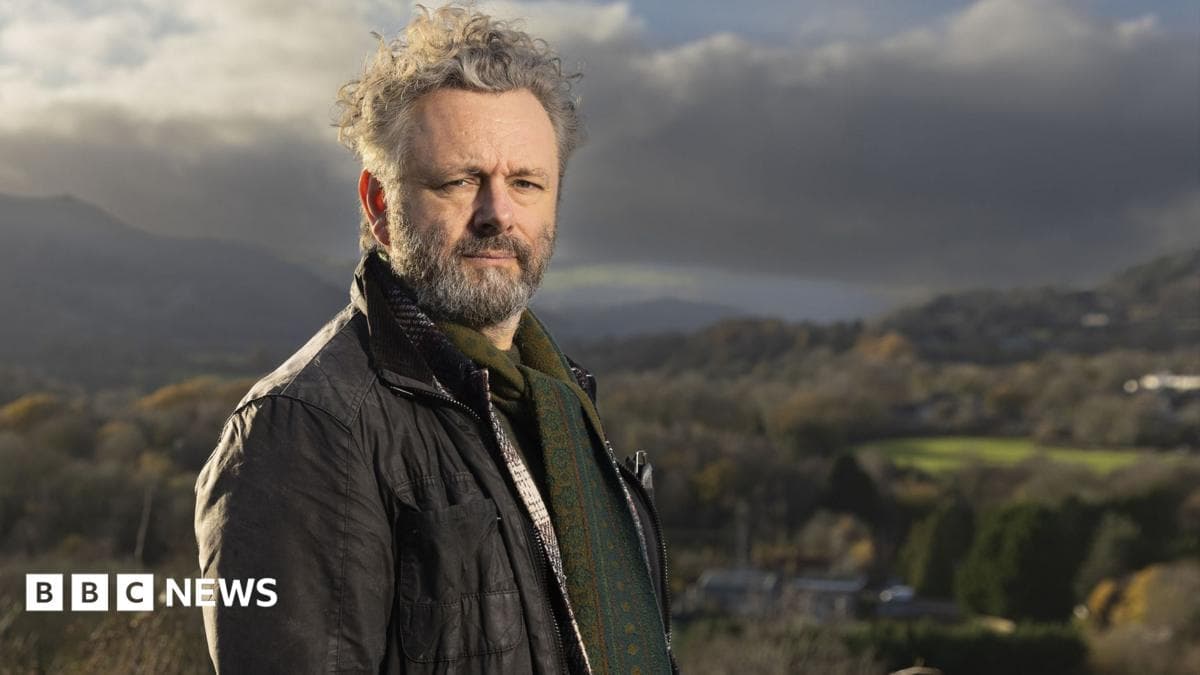 Standing on high ground above a valley, Michael Sheen looks towards the camera with rolling hills and patchwork fields stretching into the distance. He is wearing a brown jacket with a green patterned jumper. Sheen has grey hair and facial hair. Strong afternoon sunlight and heavy clouds create dramatic contrasts across the rural landscape.