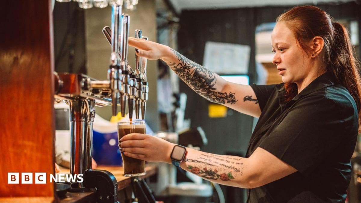 A female bartender pouring a pint of draught beer from the tap behind the bar in a busy British pub.