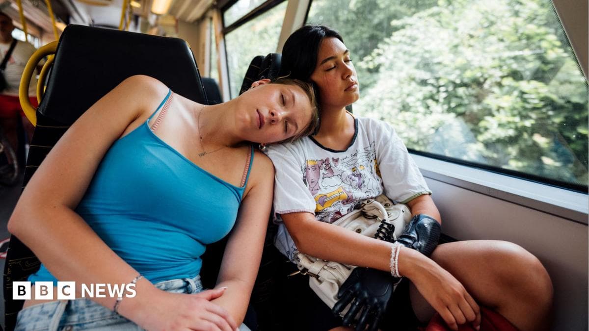 Two young women asleep on a train - one wearing a blue vest top leans on the other's shoulder