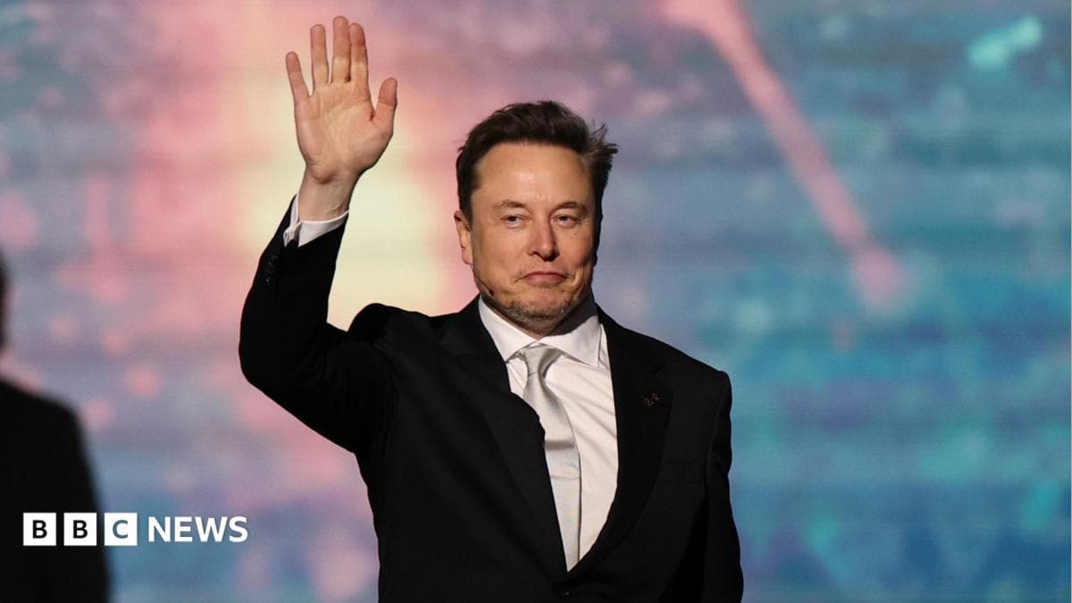 SpaceX chief executive Elon Musk walking onto a stage and waving, wearing a black suit, white collared shirt and shiny off-white neck tie.