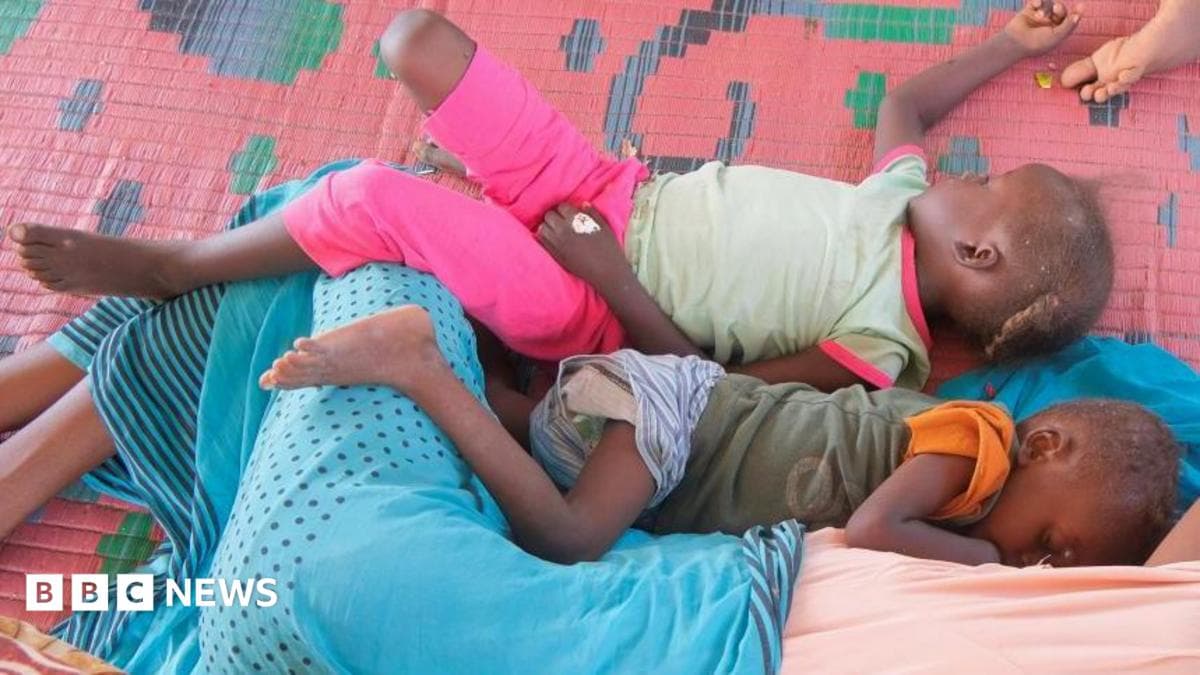 Two small children lie side-by-side on a pink grass mat next to someone covered in a cloth.
