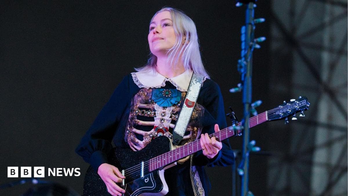 Phoebe Bridgers performs on stage holding a black guitar. She is a young woman with short, grey-coloured hair. She is wearing a black dress with a white colour and a decorated ribcage on the front of it.