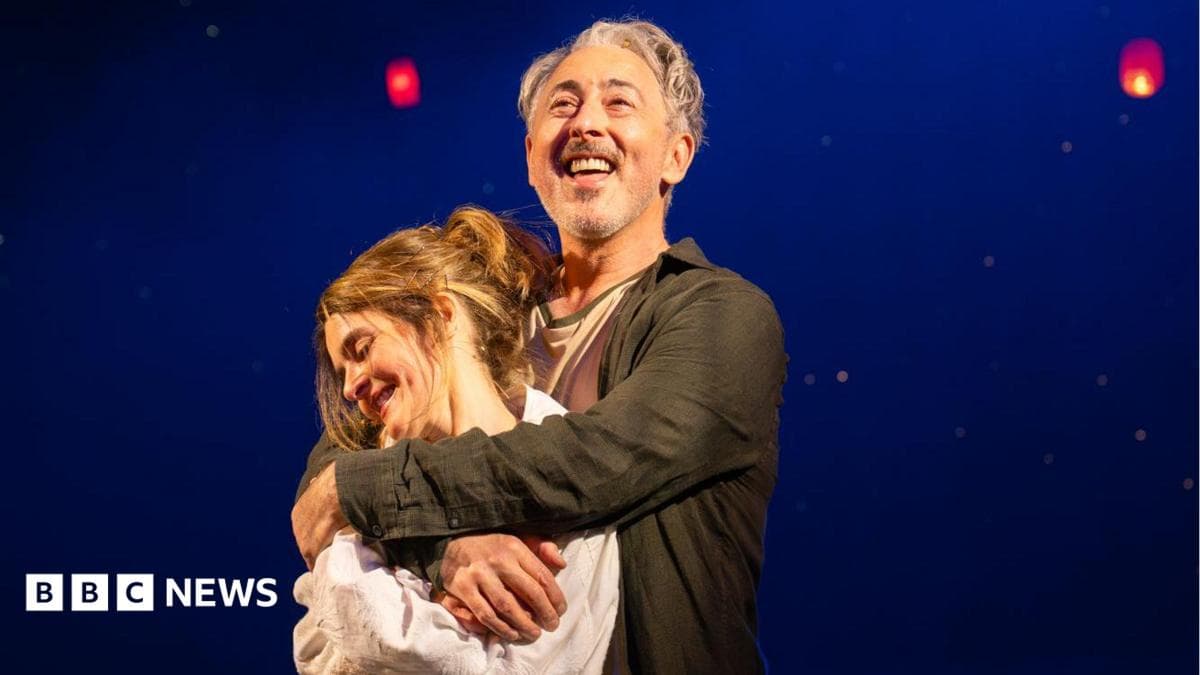 Two performers stand together on a dimly lit stage, framed by a deep blue backdrop and glowing red lanterns suspended overhead.
Warm spotlighting highlights the embrace, while the floating lights create a dreamlike, theatrical atmosphere.