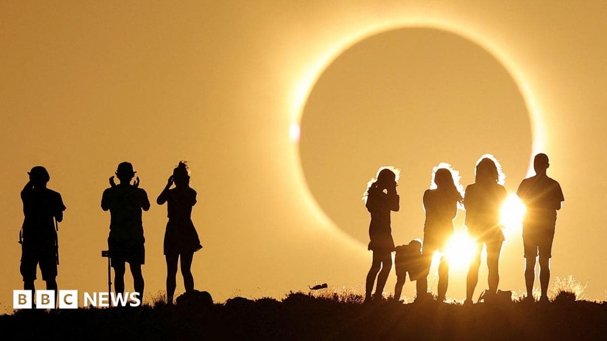 A total solar eclipse is about to occur - a darkened grey sphere appears large in the sky and bright yellow light is emanating from it drenching the sky. Two groups are gathered watching it, silhouetted against the sky