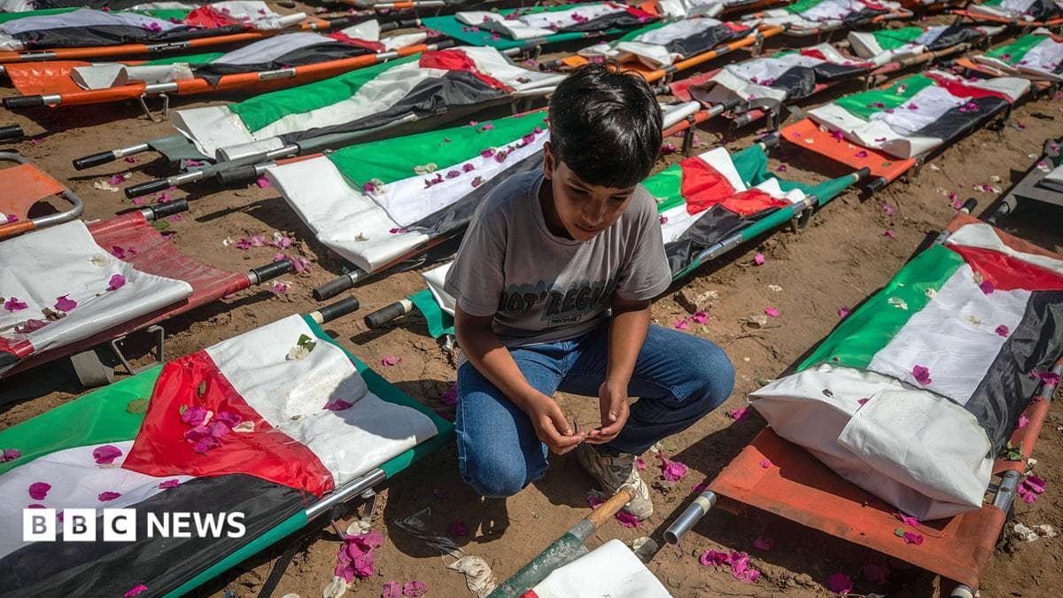 A boy crouching besides rows of stretchers draped in Palestinian flags containing the remains of 112 victims of an isreali strike in November 2023 at a Mass funerla held on 4th August 2026