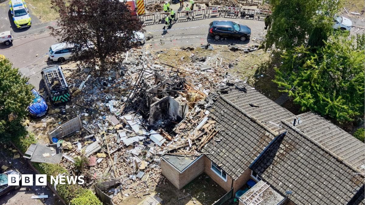 An aerial shot of three bungalows. The one on the far left is completely destroyed with just four internal walls remaining. There is debris everywhere. There is a police car parked nearby and people in high-vis vests.