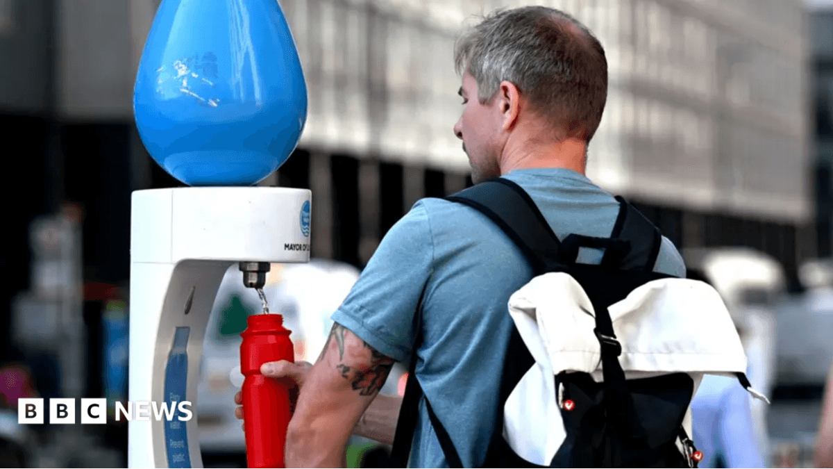 Back view of a man wearing a blue t-shirt and black backpack filling up a red water bottle at a temporary drinking fountain