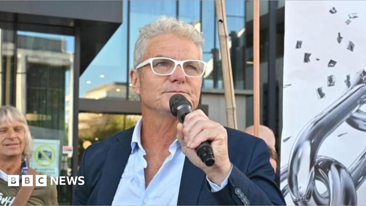 A man wearing white-rimmed glasses and a dark suit, holds a microphone