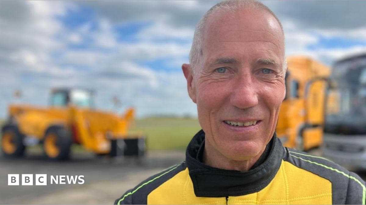 A man looks towards the camera wearing a yellow and black driving suit with the "JCB" logo on the front.