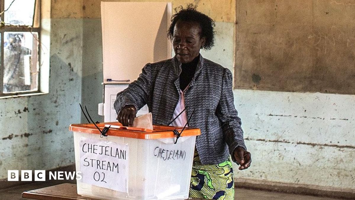 A woman in a patterned jacket and African wrap for a skirt stands at a ballot box putting her vote in. She is in an empty classroom and a blackboard can be seen behind her.