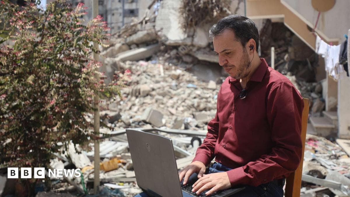 A Palestinian man works on a laptop on a chair outside, surrounded by rubble, in Gaza