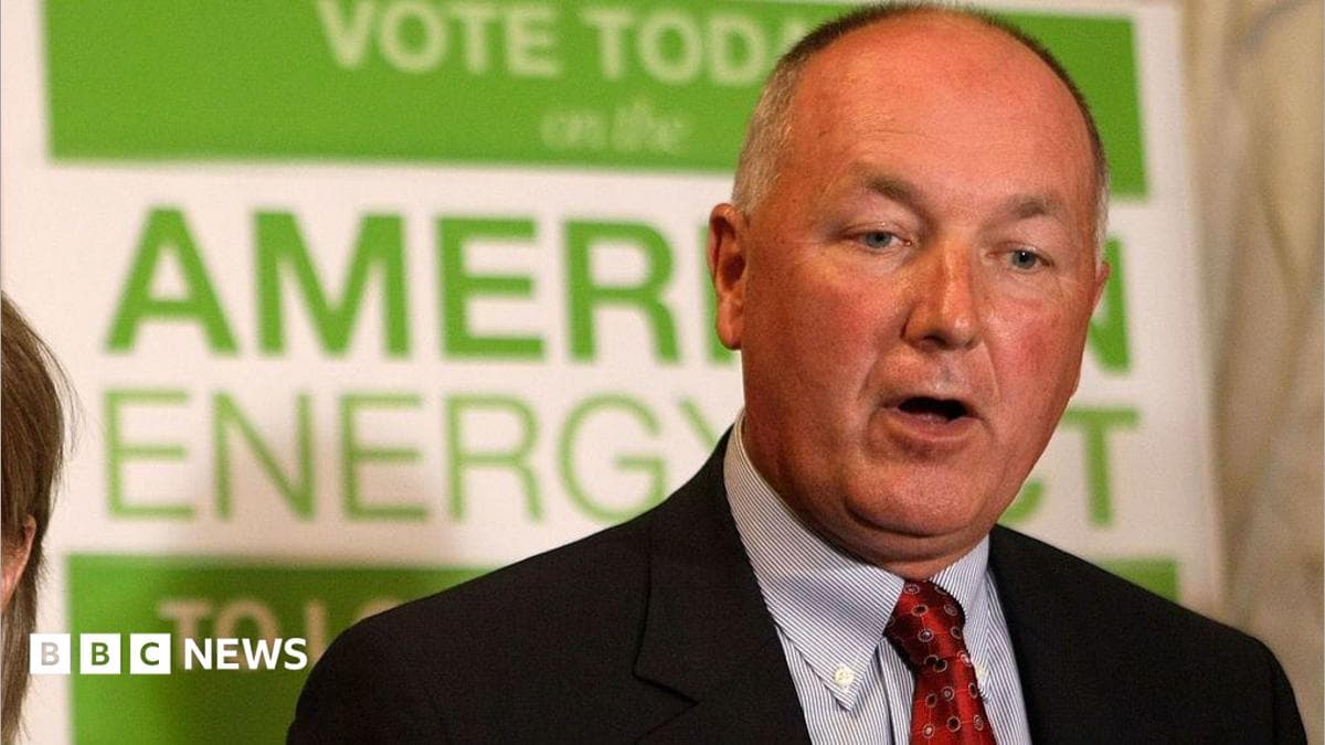 Pete Hoekstra, with short grey hair, and in a dark suit coat, red tie and patterned blue shirt, stands in from of a green and white campaign sign promoting American energy.