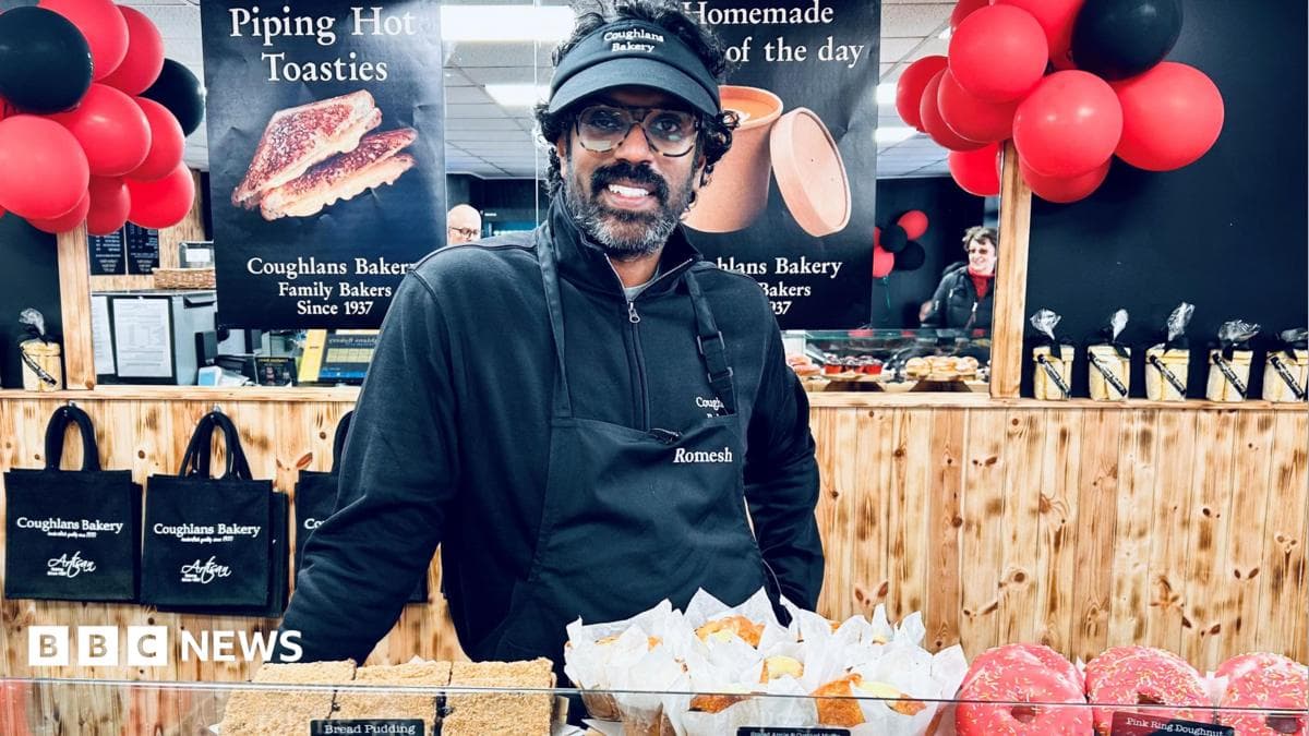 Romesh stood behind a counter of baked goods. He is wearing an apron and black cap and is smiling directly at the camera. There are red and black balloons behind him