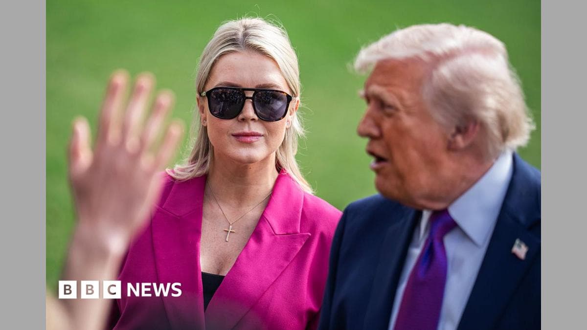 Karoline Leavitt wears sunglasses and a pink blazer as she stands behind Donald Trump during a press gaggle.