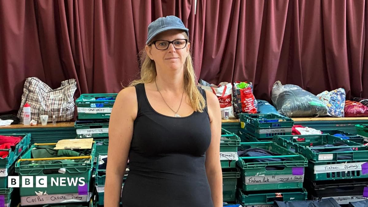 Zia Lee stands in a black vest top with a denim cap on and black glasses and long blonde hair. Behind her are green crates with school names and carefully folded uniforms in them.