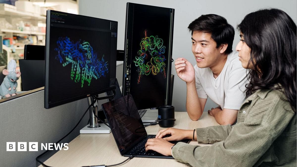 Two people working together at a desk in what appears to be a laboratory or research office environment. Large computer monitors display colorful three-dimensional molecular or protein structure models against dark backgrounds, while a laptop on the desk shows lines of code or data. One person is seated using the laptop, and another is leaning toward the screens as both focus on the scientific visualizations. Laboratory shelving and equipment are visible in the background, suggesting a biomedical, biotechnology, or computational research setting. The scene emphasizes the use of computer-based analysis and visualization tools to study complex biological structures.