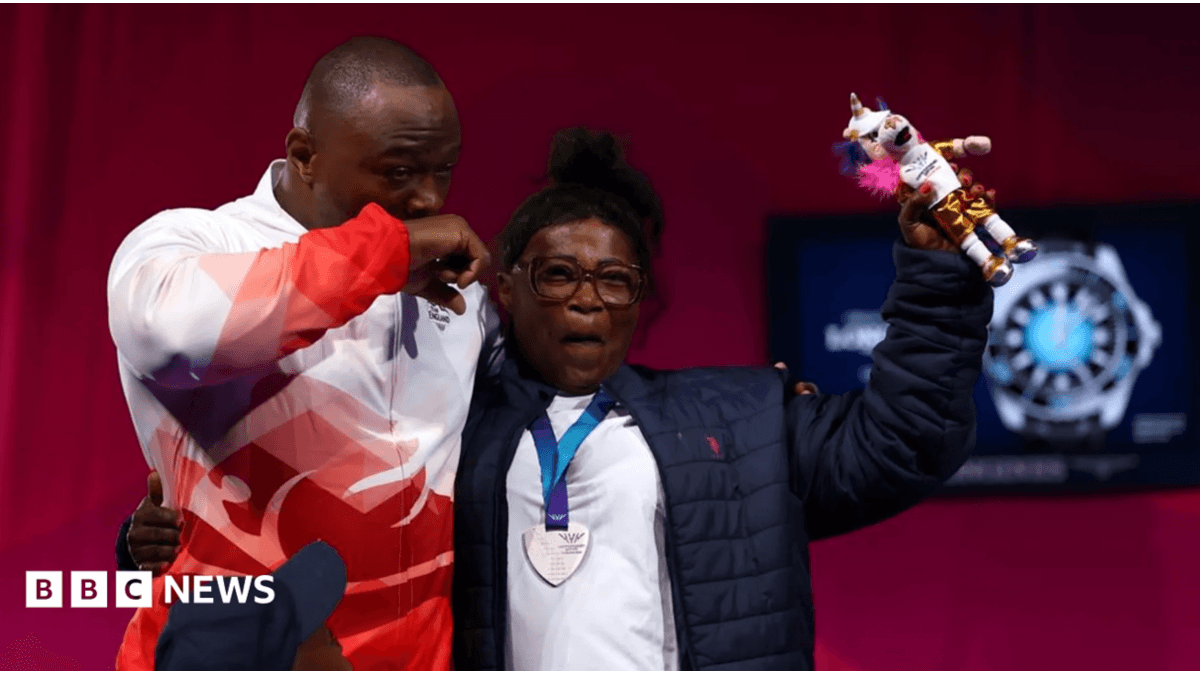 Person wearing glasses, a dark quilted jacket and a silver medal on a blue ribbon stands beside another person in a white and red sports jacket. The medal wearer raises a colourful mascot toy while standing on a sports event stage with a red backdrop.