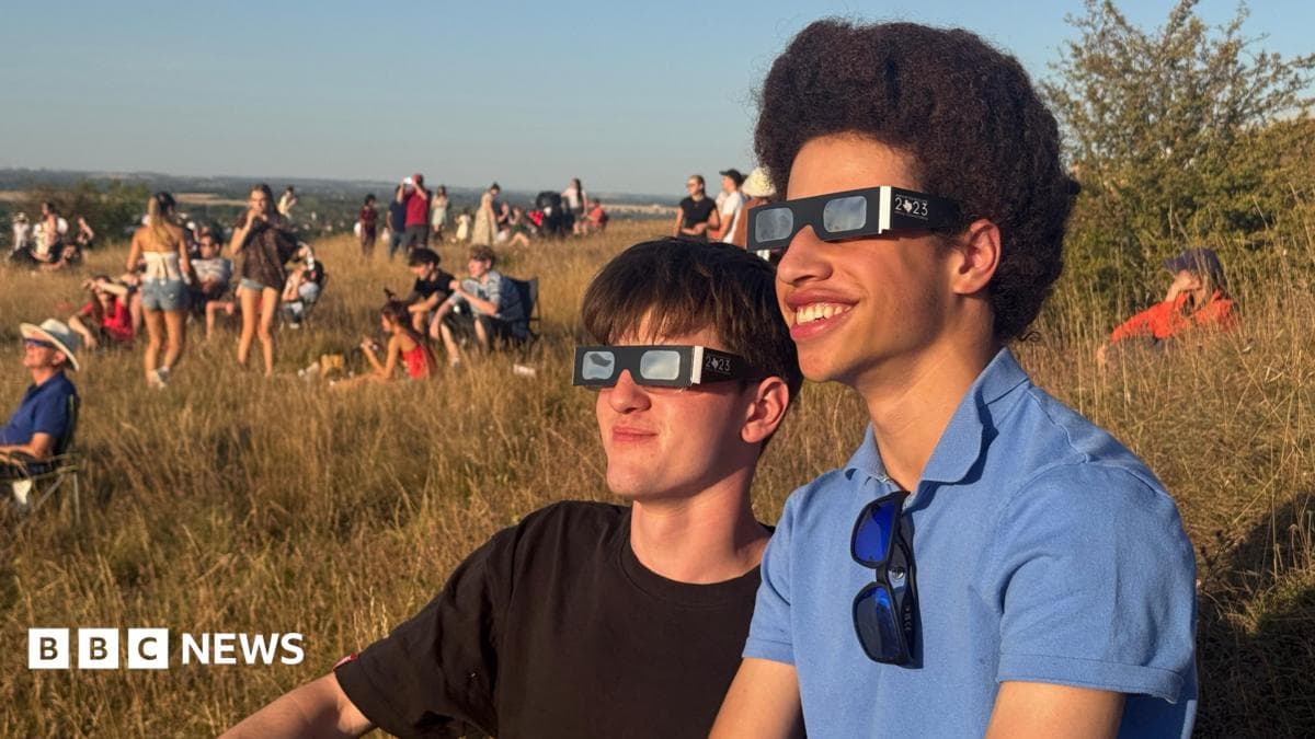 Two young men wearing eclipse glasses sit on a grassy hillside while crowds gather in the background to watch a partial solar eclipse.