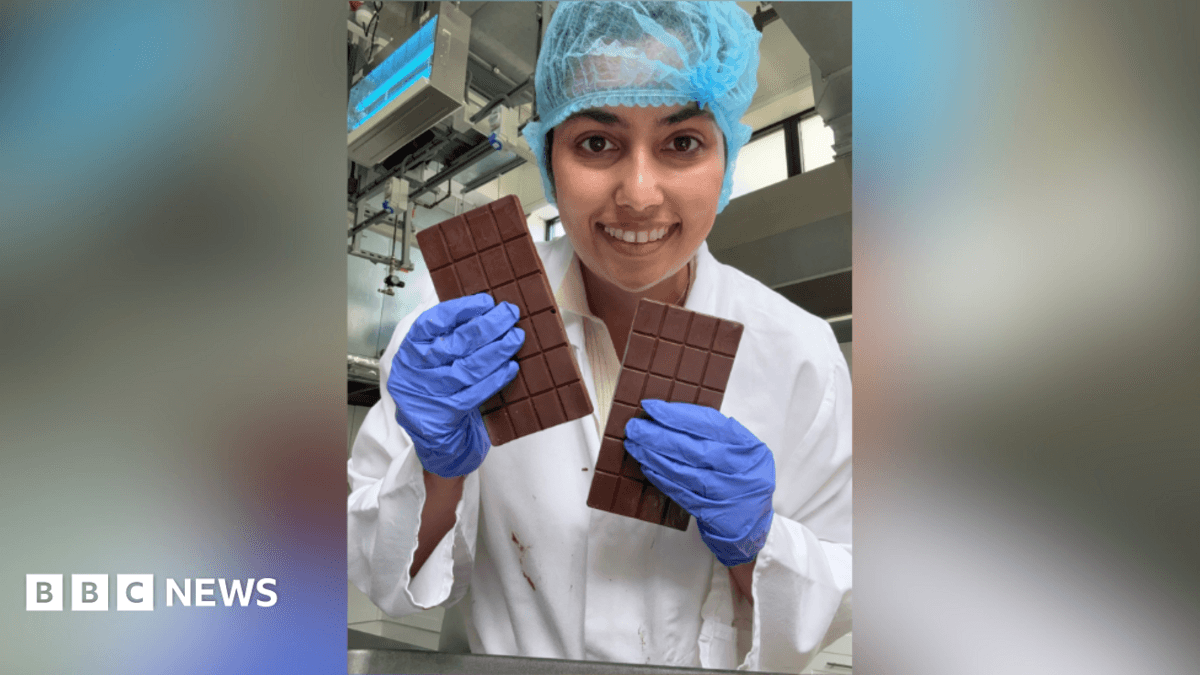 A woman holding two bars of chocolate while smiling into the camera. She is wearing a white gown with purple gloves and a blue hair net.