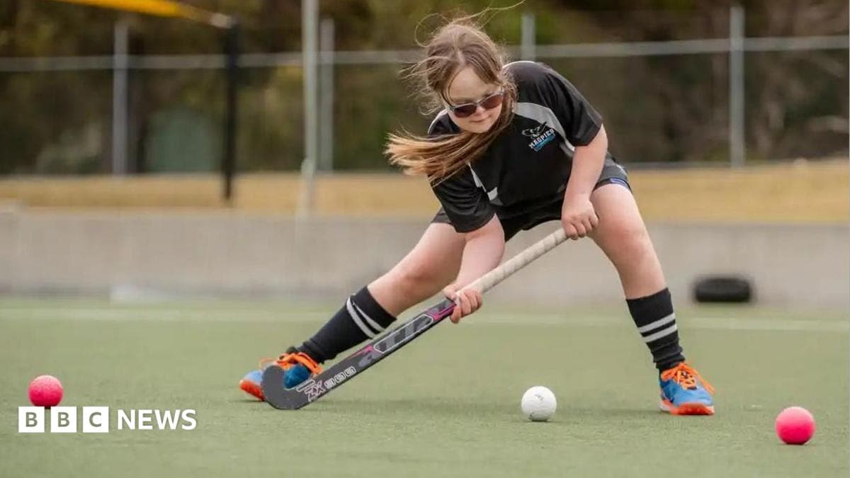 A girl wearing black sports shorts and t-shirt leans forward to hit a white ball with a hockey stick