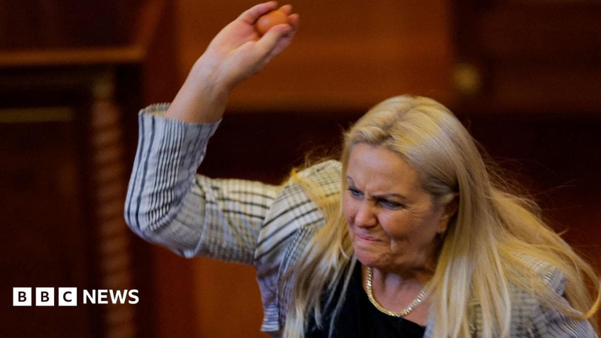 Blond woman wearing white and black blazer lifting right arm and holding egg