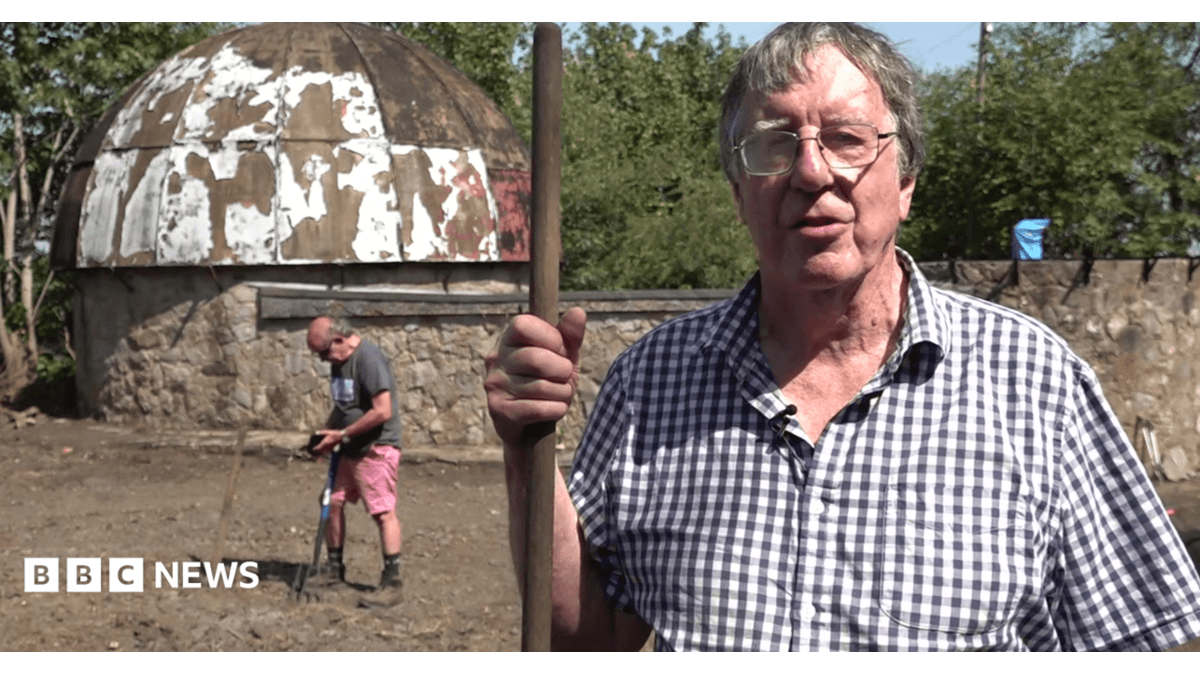 Miles is stood at the observatory site with a man working on the ground in the background. He is wearing a checked shirt and glasses and is holding a metal pole. A very rundown and dirty dome can be seen behind him.