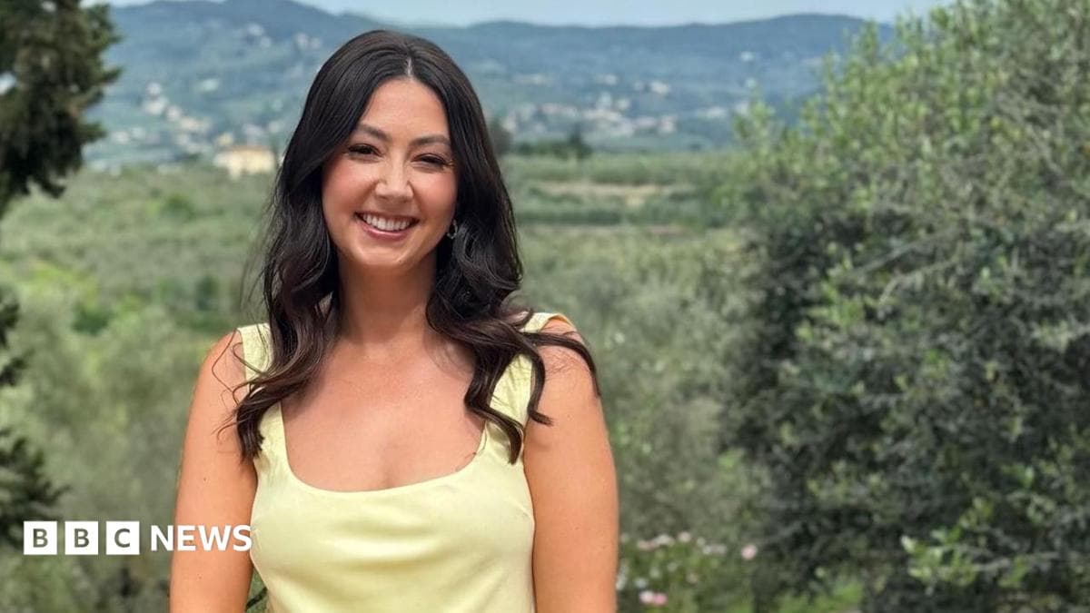 A woman with long dark hair and a yellow sleeveless top smiling in front of a countryside background.