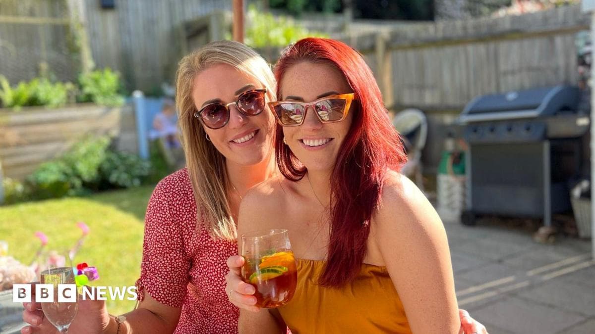 Friends Sarah (left) and Leanne (right) wearing sunglasses, smiling and holding drinks as they sit in a garden.