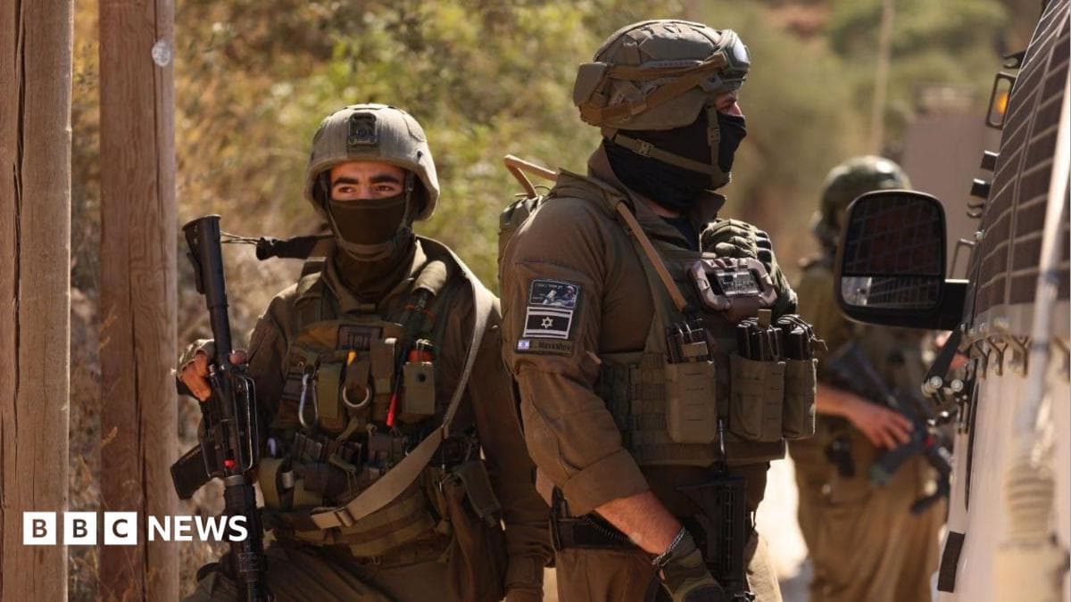 Two IDF soldiers in military gear and face coverings block a road near the West Bank village of Qusra near Nablus. One soldier is seen facing towards the side of a van while a second holding a rifle is facing towards the camera but looking off in the distance.