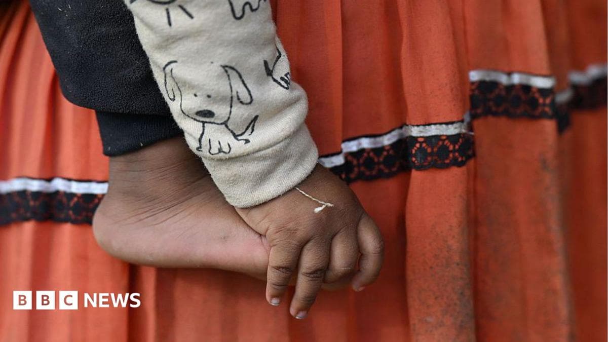 A close-up of a small child’s bare foot and hand is shown against a backdrop of their mother's skirt - a traditional indigenous outfit. It appears the child is being held by their mother. The child is wearing a light-coloured sleeve printed with a simple cartoon dog, and the hand hangs loosely beside the foot.