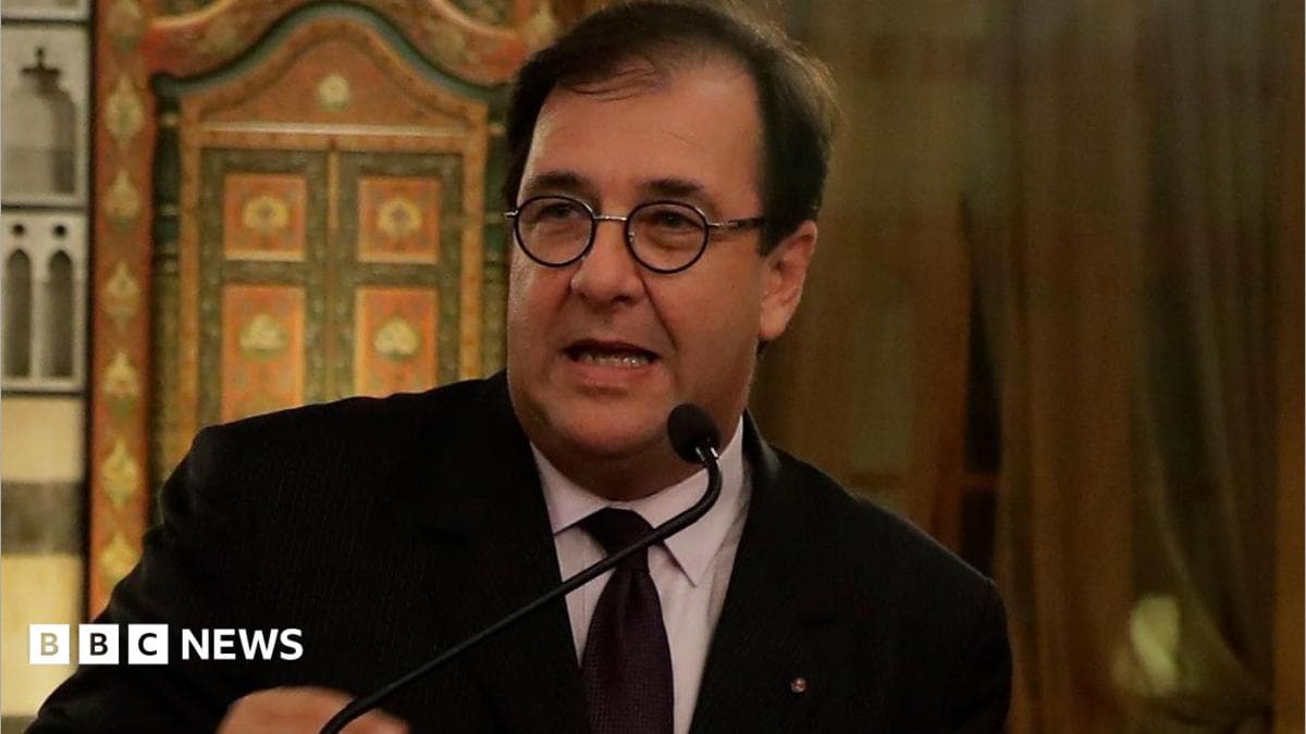 Bruno Foucher in a black suit and tie and a white shirt speaks to a microphone