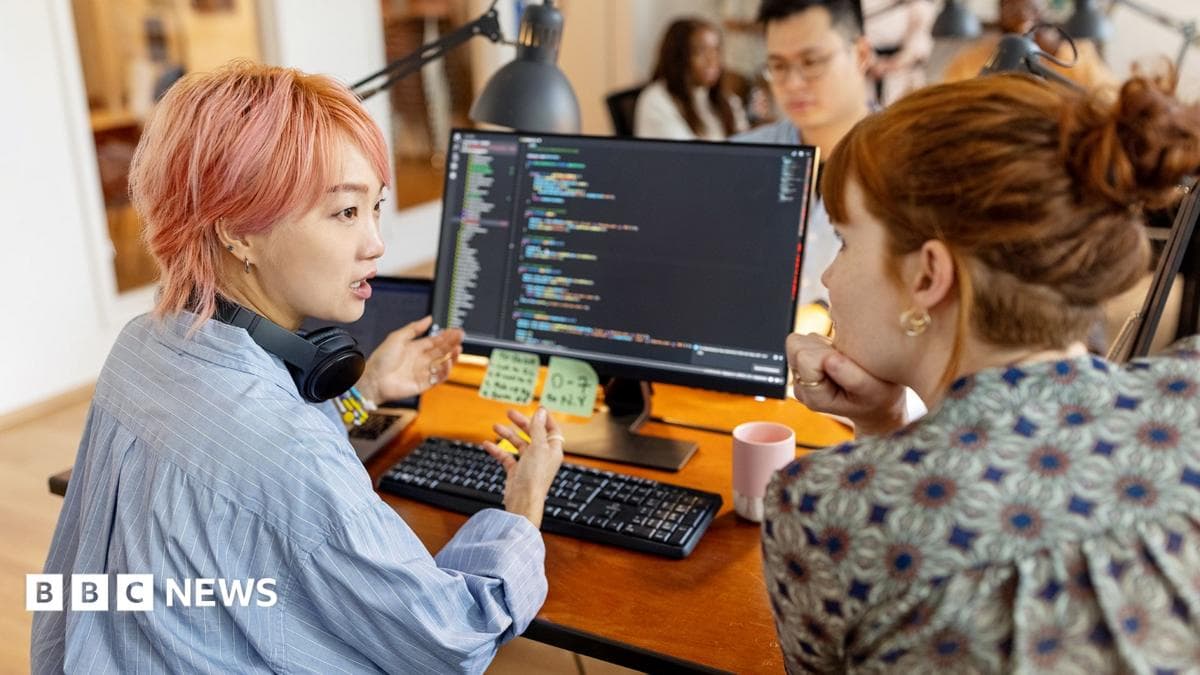 Stock photo shows a tech worker with pink hair sits at a desk and gestures while talking to a colleague who rests their chin on their hand, with other workers in the background, in an office environment with coding language on the computer screen in view.