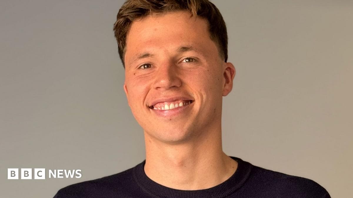 Freddie Woodward: a young man with short dark hair sits on a grey sofa, smiling at the camera. He has short brown hair and wears a navy jumper.