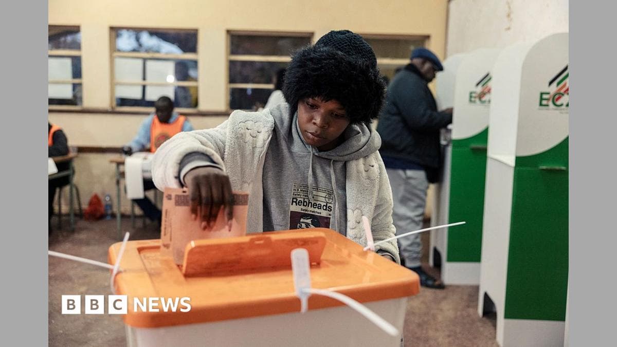 A woman in a black woolly hat and thick white cardigan places a ballot paper in a plastic ballot box with an orange lid. Some polling booths and election officials can be seen behind her.