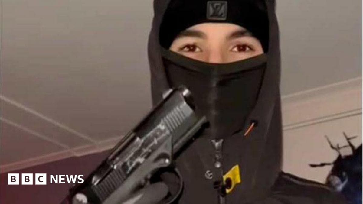 A teenager wearing a black balaclava and hoodie holding up a handgun, with his gloved hand on the trigger