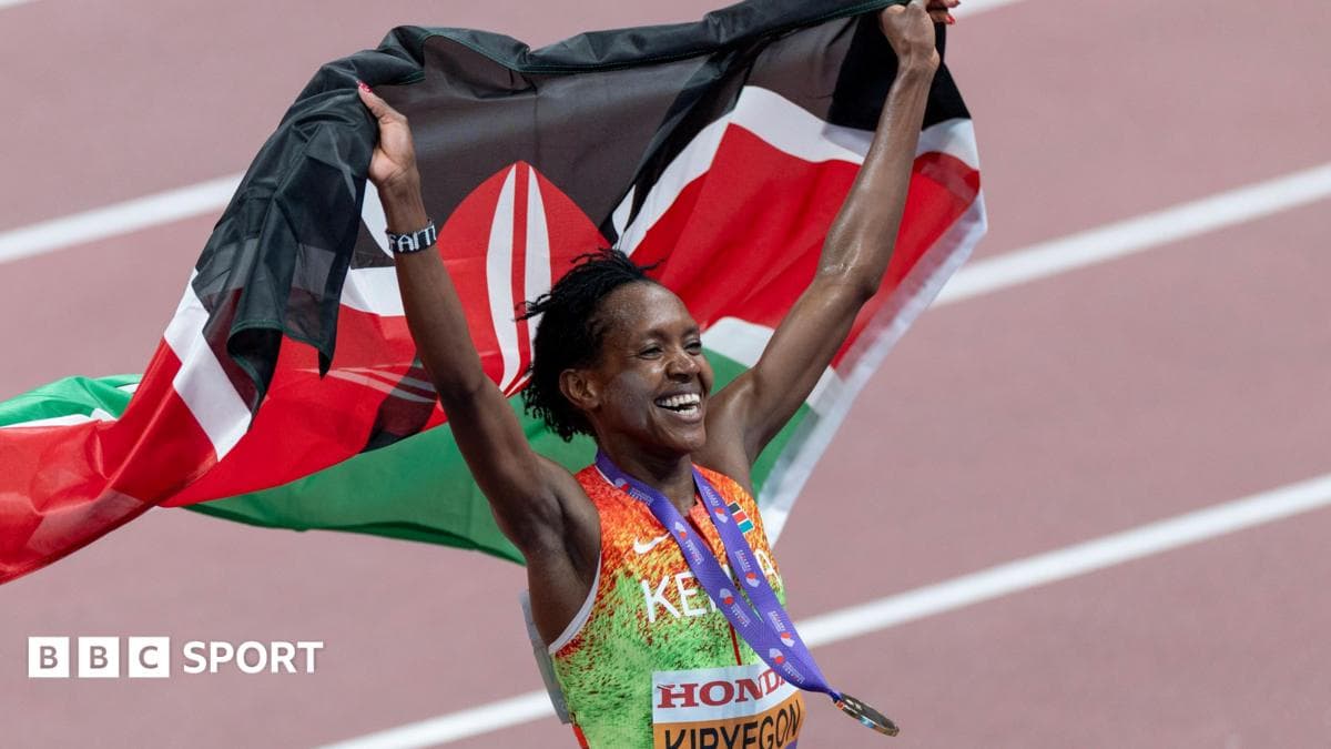 Faith Kipyegon, wearing an orange and green Kenyan athletics vest, with a gold medal on a purple ribbon bouncing around her neck, smiles broadly as she holds a Kenyan flag aloft behind her. The runner is visible from stomach up while a terracotta-coloured athletics track with white lines is seen out of focus in the background