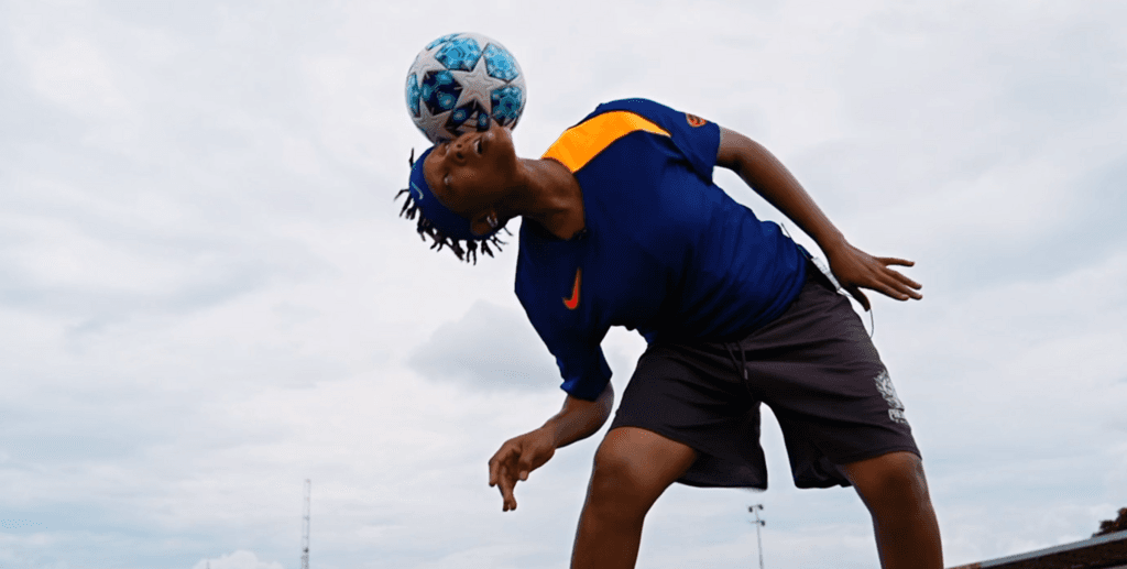 A camera shoots up from a low level to show a female freestyler leaning to the side while balancing a football on her head underneath a cloudy sky